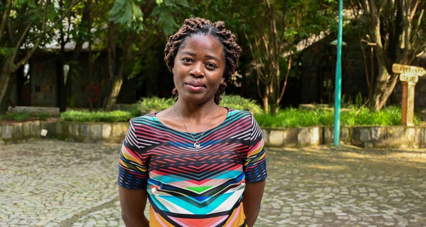 A woman stands in a tree-lined courtyard, looking at the camera with a slight smile. She wears a colourful, geometric-patterned dress and a pendant necklace.