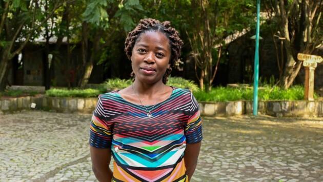 A woman stands in a tree-lined courtyard, looking at the camera with a slight smile. She wears a colourful, geometric-patterned dress and a pendant necklace.